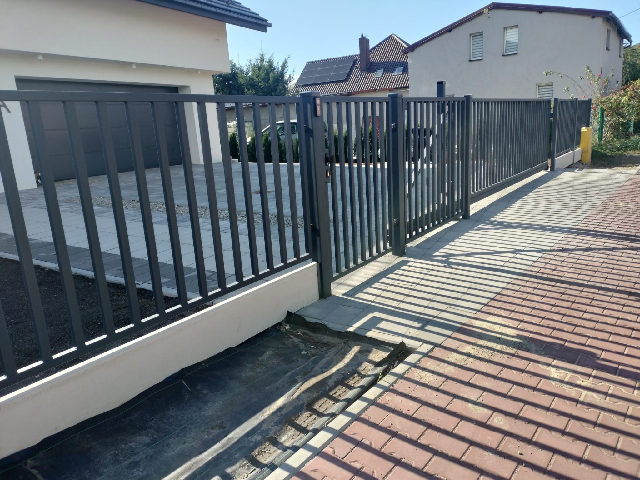 Nowoczesne, grafitowe ogrodzenie posesji z przesuwną bramą wjazdową w Mysłowicach, z widocznymi cieniami rzucanymi przez pionowe elementy konstrukcji na chodnik z czerwonej kostki.