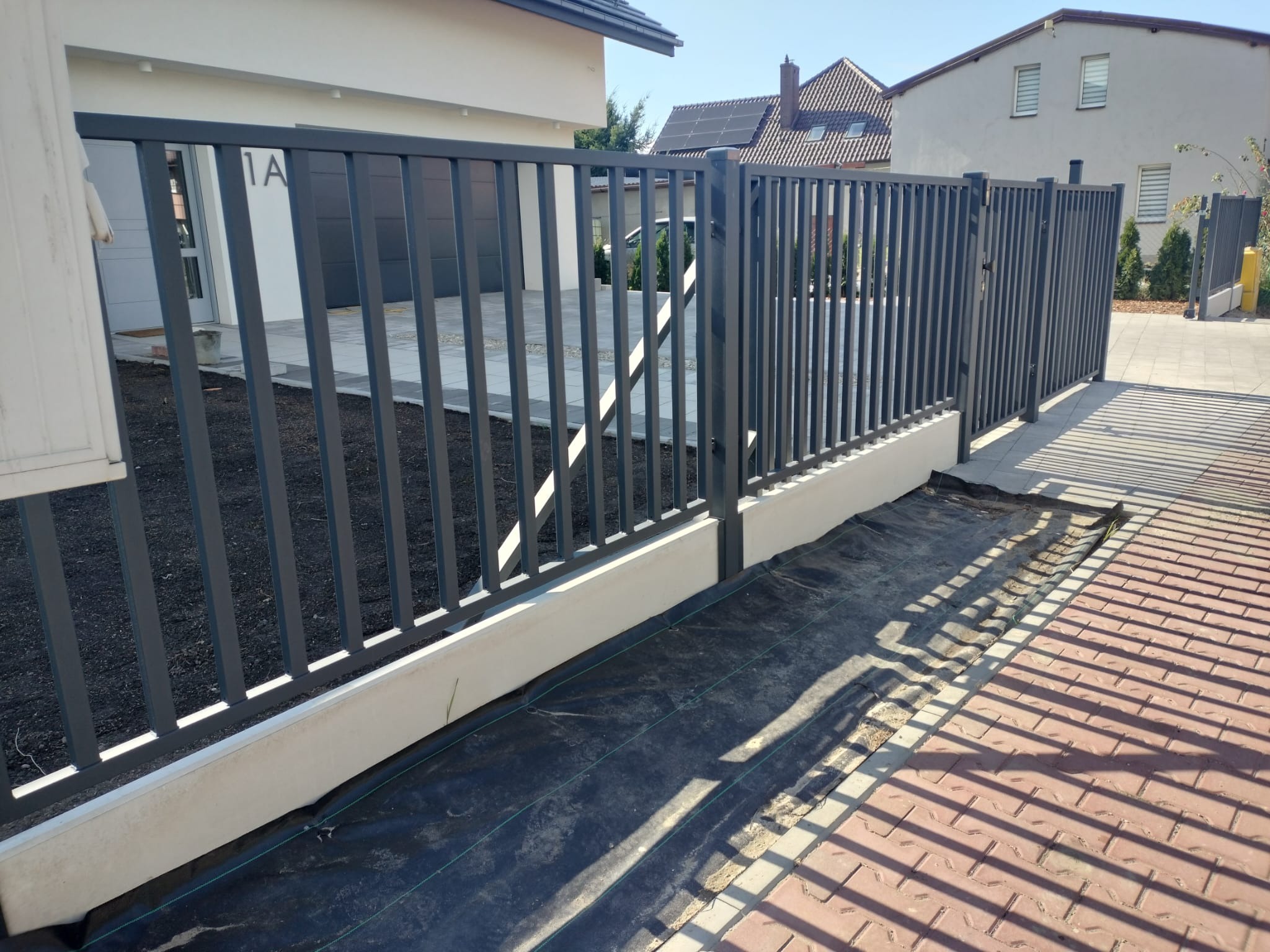 Nowoczesne, metalowe ogrodzenie panelowe w kolorze grafitowym, zamontowane na niskiej, betonowej podmurówce, z widoczną czarną agrowłókniną wzdłuż ogrodzenia i kostką brukową na podjeździe.