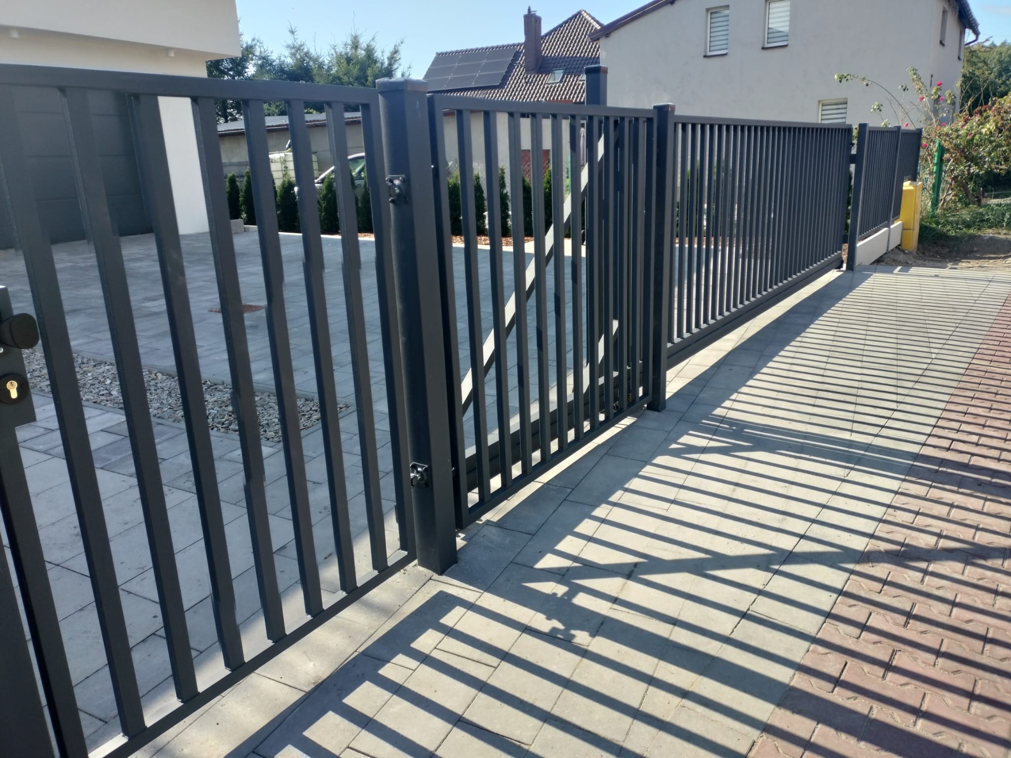 Nowoczesna, przesuwna brama wjazdowa z pionowych, metalowych profili w kolorze grafitowym, rzucająca regularny cień na brukowany podjazd.