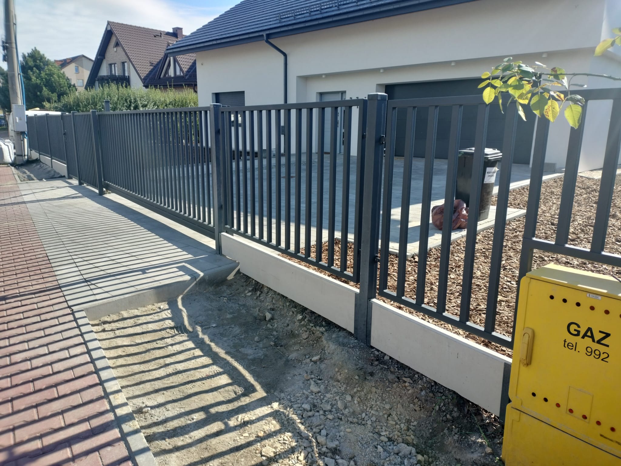 Nowoczesne ogrodzenie metalowe w kolorze grafitowym z pionowymi prętami, zamontowane na betonowej podmurówce, widoczne na tle jasnego domu jednorodzinnego i chodnika z kostki.