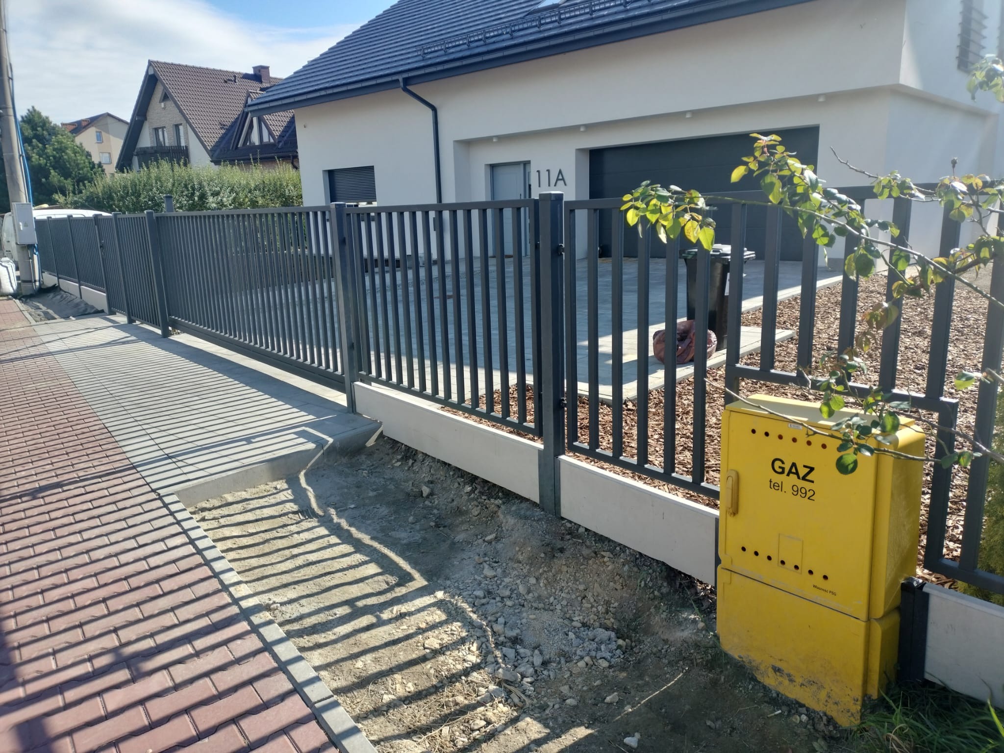 Nowoczesne ogrodzenie metalowe w kolorze grafitowym, z pionowymi prętami, otaczające posesję z domem jednorodzinnym; widoczna skrzynka gazowa w kolorze żółtym i fragment chodnika z kostki.