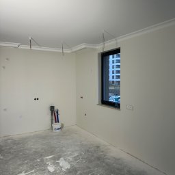 Wykończenia Wnętrz Mateusz Cirocki - Pokój w trakcie remontu z widocznymi śladami po pracach na podłodze, świeżo pomalowane ściany w jasnym odcieniu, dekoracyjny gzyms sufitowy, okno z widokiem na budynek mieszkalny, przygotowane...