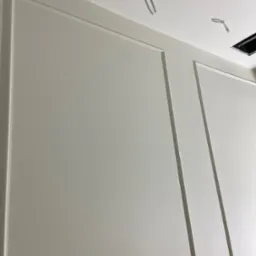 Pomalowana na biało ściana z dekoracyjnymi listwami w stylu klasycznym, widoczne wystające przewody elektryczne i otwór w suficie.