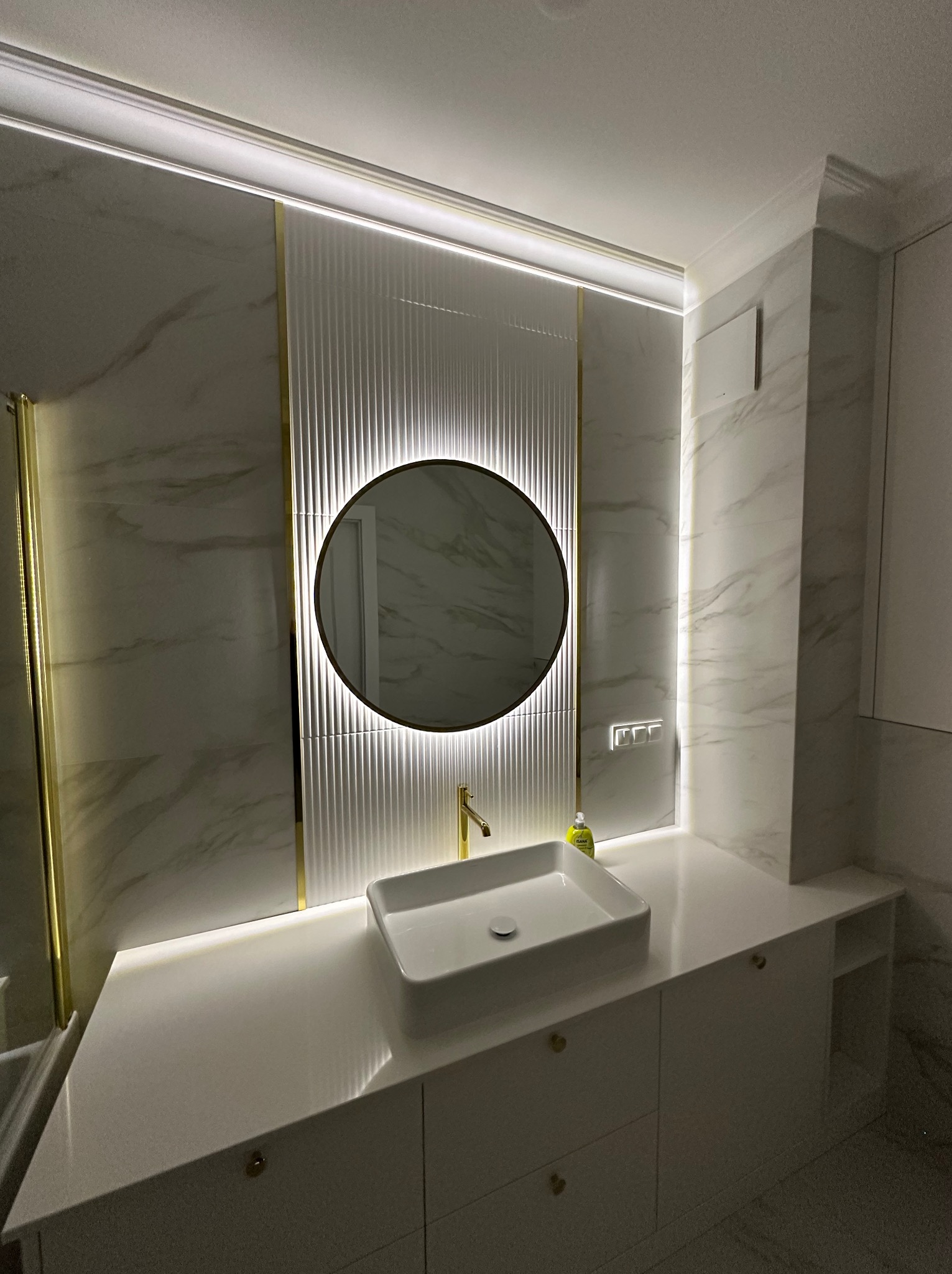 Nowoczesna łazienka z umywalką nablatową, okrągłym lustrem podświetlanym LED, marmurowymi płytkami z elementami złota i oświetleniem podszafkowym.