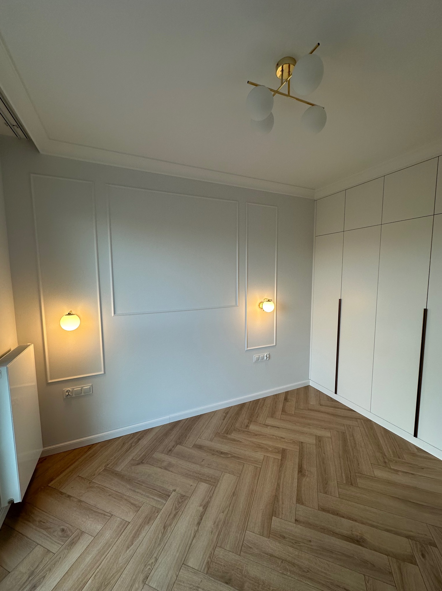 Pokój po remoncie: parkiet w jodełkę, białe ściany z dekoracyjnymi listwami, złote kinkiety z kulistymi kloszami, biała szafa w zabudowie z długimi, ciemnymi uchwytami.