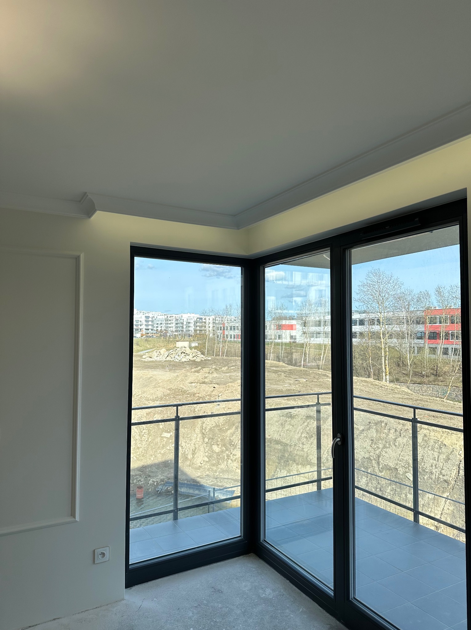 Wnętrze pokoju z dużym oknem balkonowym i ozdobną listwą przysufitową, widok na plac budowy zza balkonu.