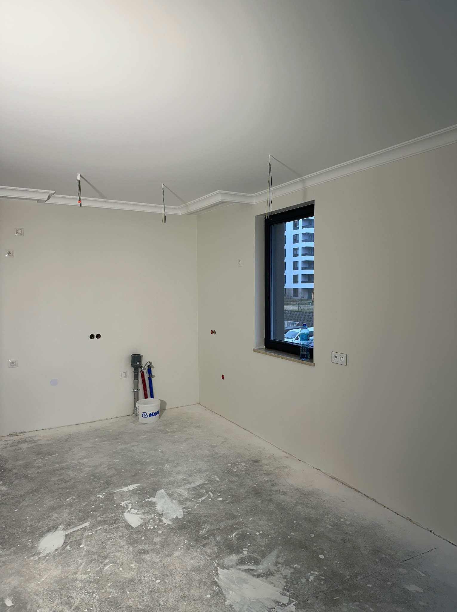 Pokój w trakcie remontu z widocznymi śladami po pracach na podłodze, świeżo pomalowane ściany w jasnym odcieniu, dekoracyjny gzyms sufitowy, okno z widokiem na budynek mieszkalny, przygotowane...