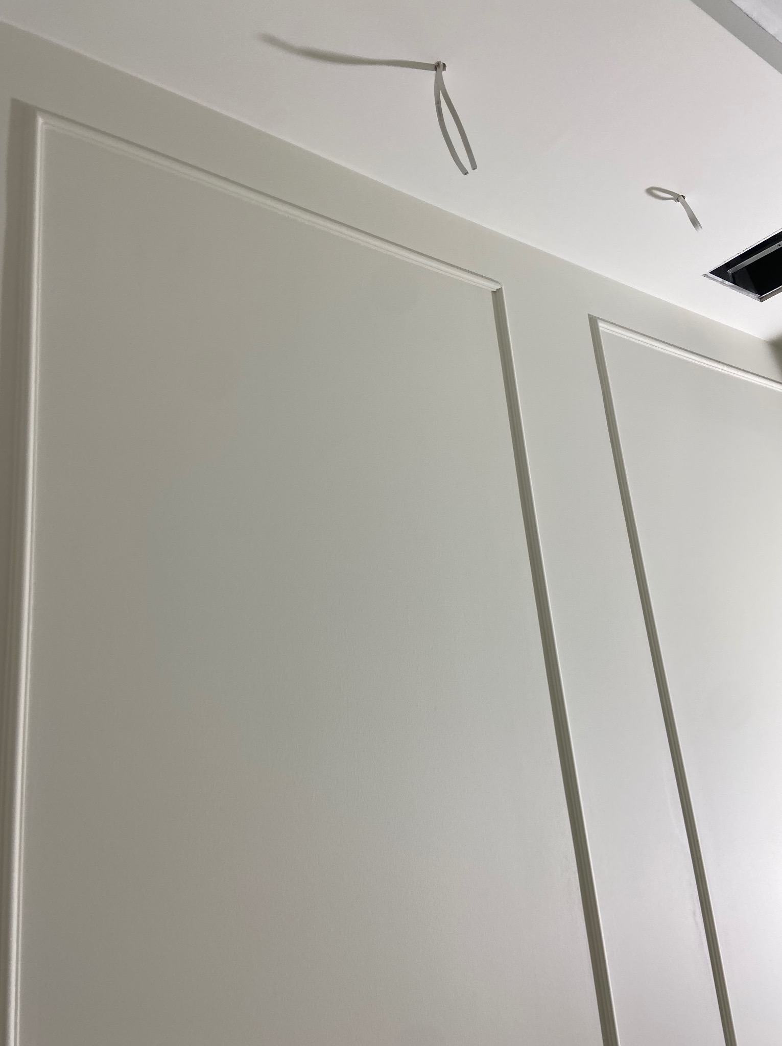 Pomalowana na biało ściana z dekoracyjnymi listwami w stylu klasycznym, widoczne wystające przewody elektryczne i otwór w suficie.