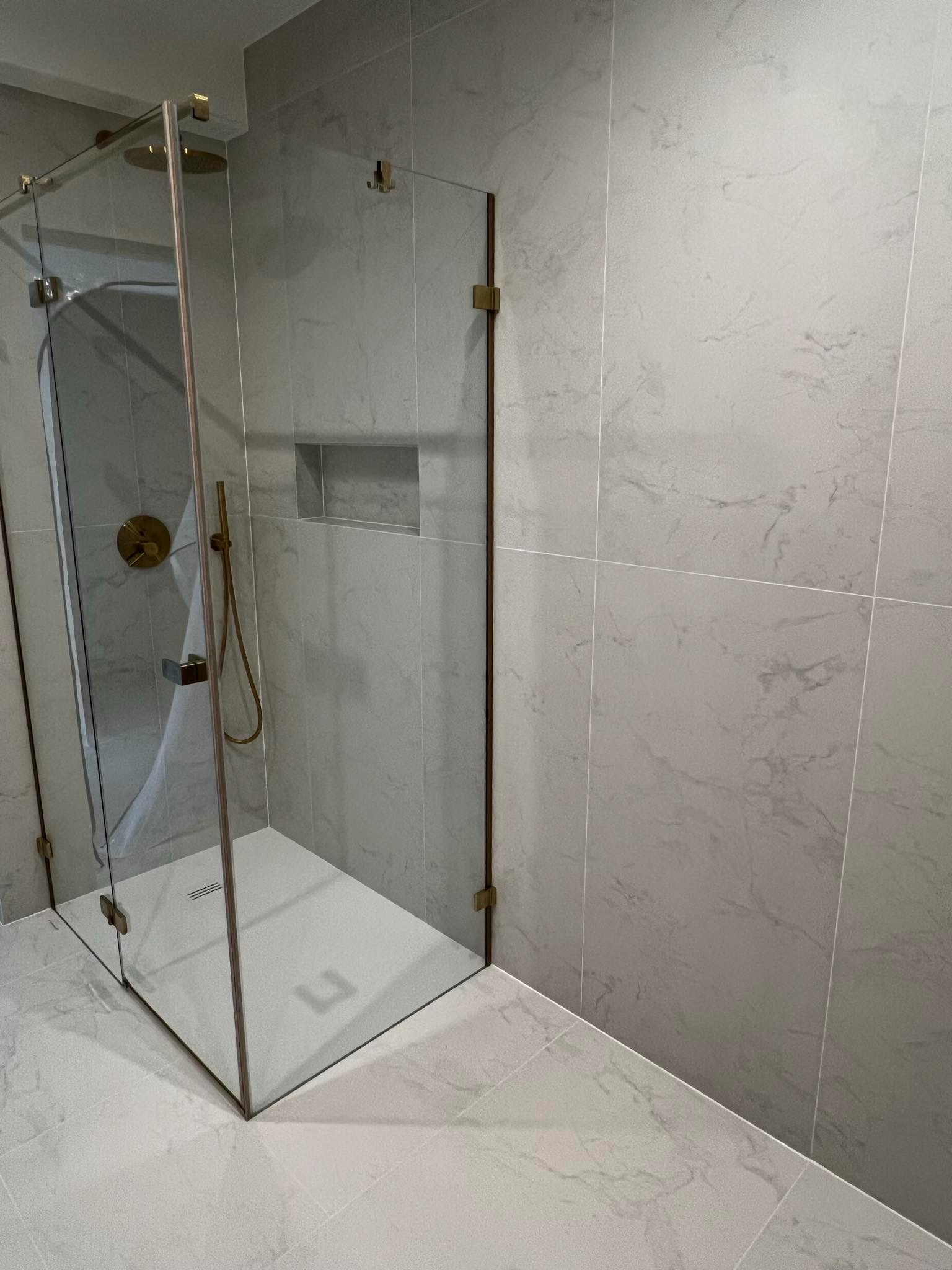 Nowoczesna kabina prysznicowa z elementami w kolorze mosiądzu, szklane drzwi uchylne, płytki imitujące marmur na ścianach i podłodze, wnęka prysznicowa w ścianie.