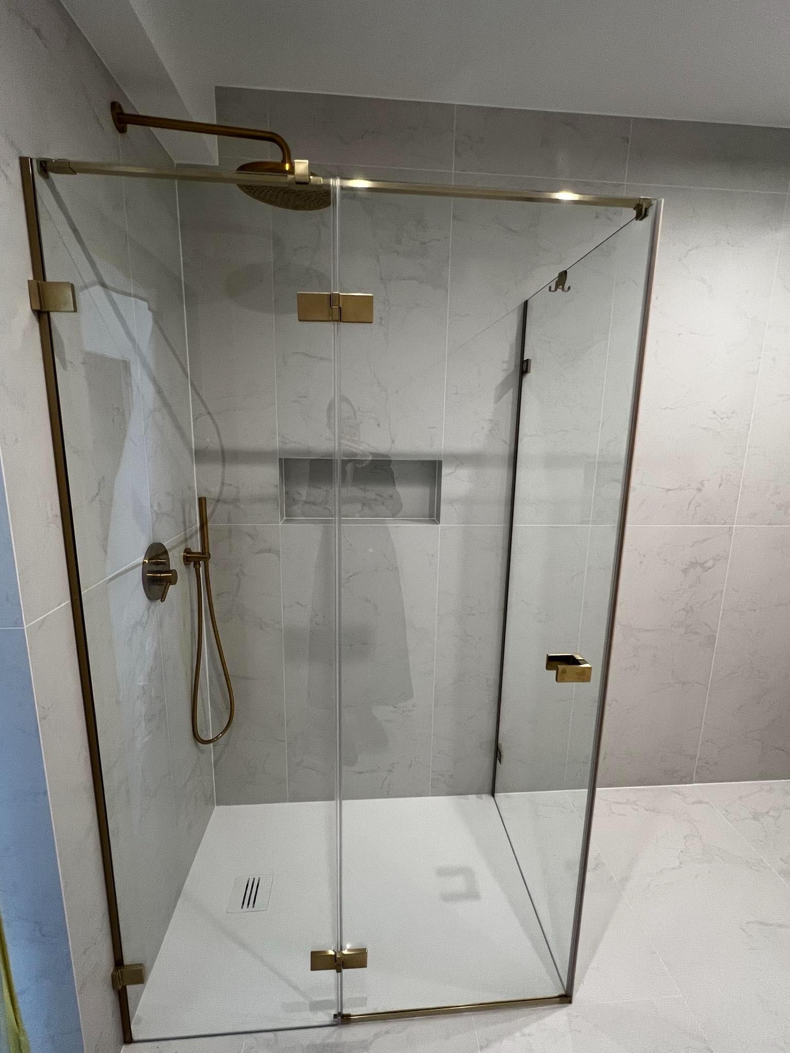 Nowoczesna kabina prysznicowa z elementami w kolorze szczotkowanego złota, szklane drzwi z minimalistycznymi zawiasami, jasne płytki imitujące marmur na ścianach, wbudowana półka w ścianie, odpływ...