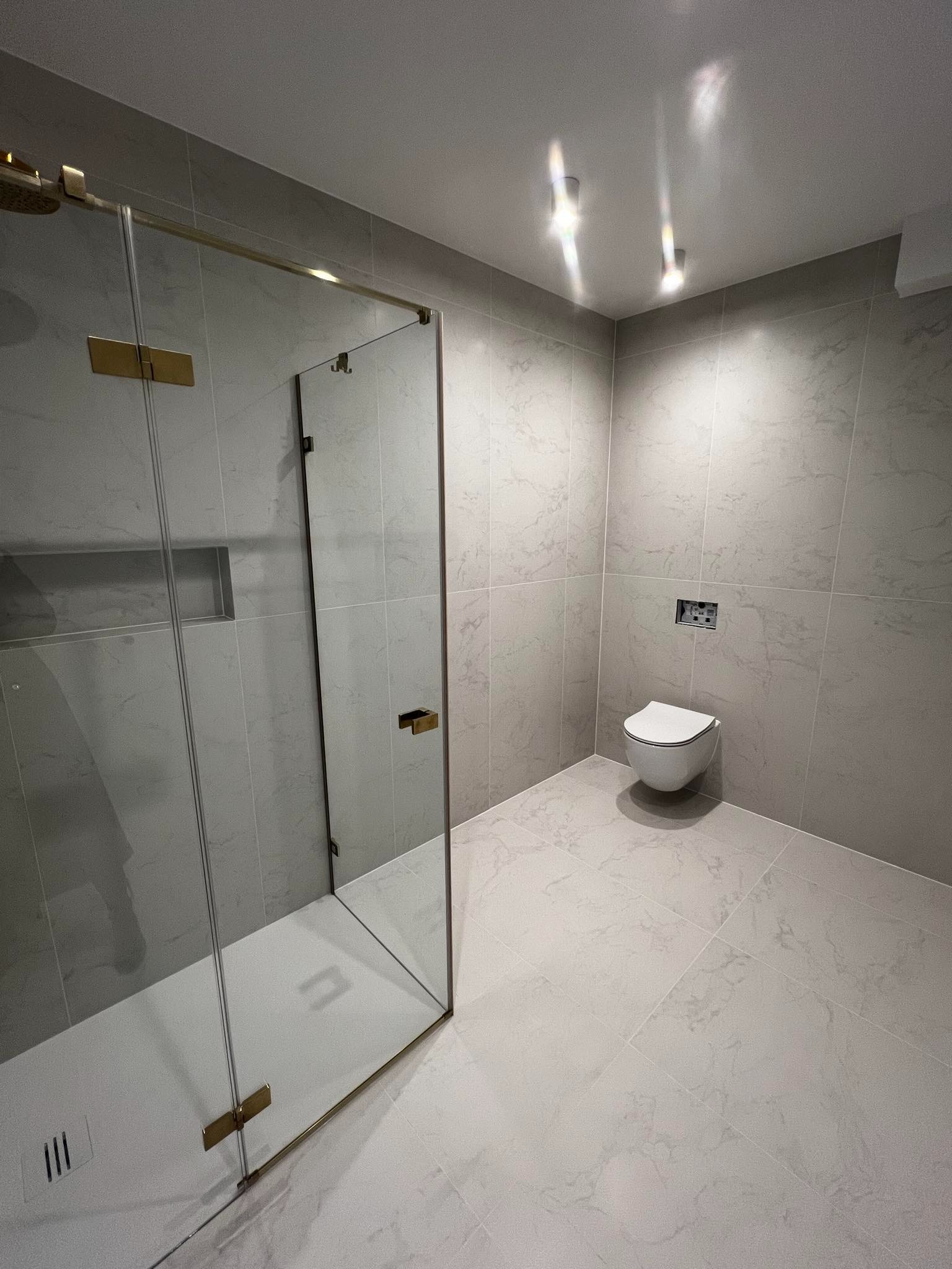 Nowoczesna łazienka z szarymi płytkami imitującymi marmur, kabina prysznicowa ze złotymi detalami i podwieszana toaleta.