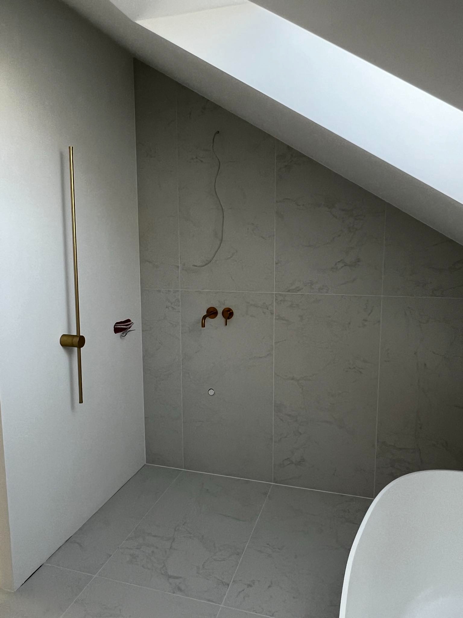 Łazienka na poddaszu w trakcie wykańczania; widoczne szare płytki imitujące marmur, złota bateria prysznicowa i miedziane krany, instalacja elektryczna wystająca ze ściany, fragment wanny.