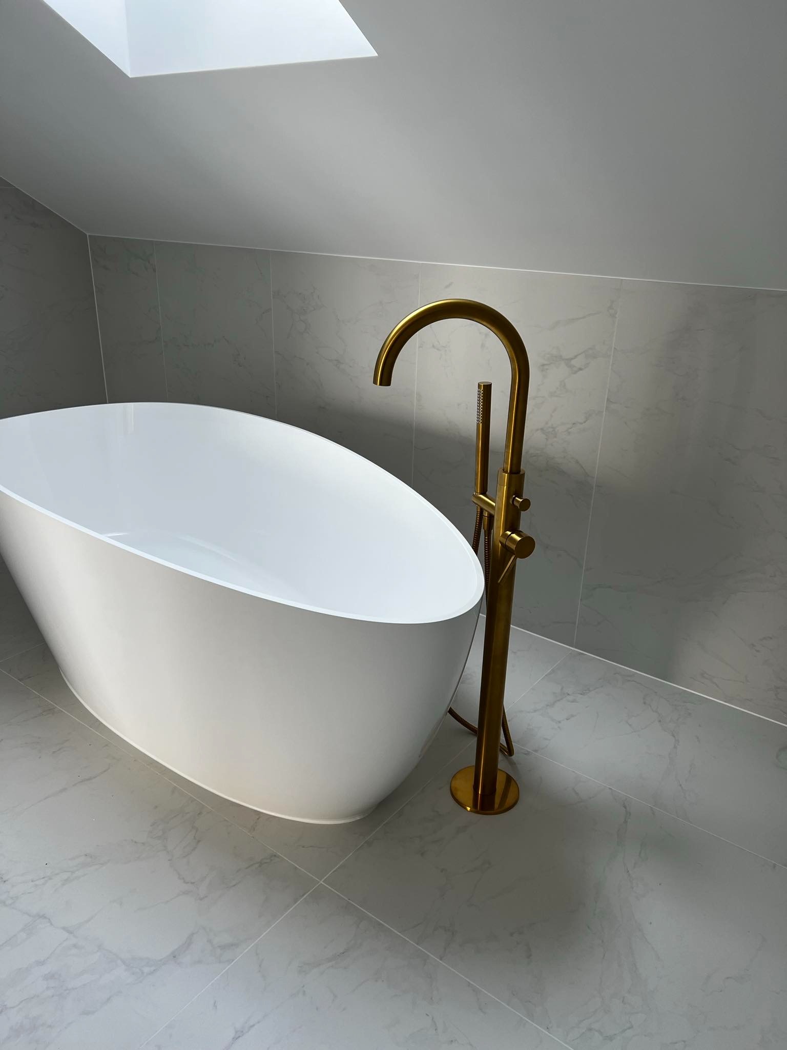 Wolnostojąca biała wanna z złotą baterią pod skosem dachu z oknem, wykończona szarymi płytkami imitującymi marmur.