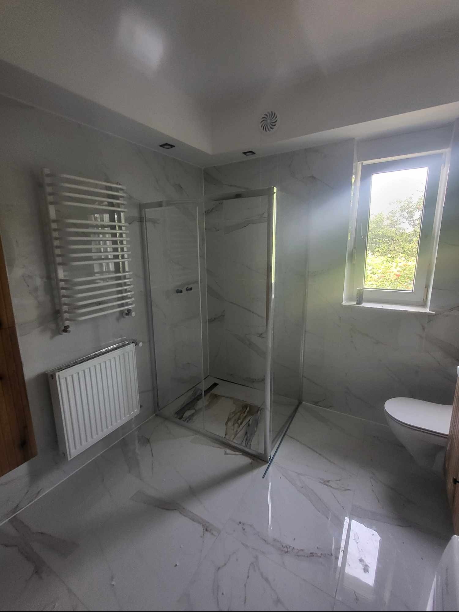 Nowoczesna łazienka z marmurowymi płytkami, kabiną prysznicową ze srebrnymi profilami, grzejnikiem drabinkowym i standardowym grzejnikiem płytowym, widok na zewnątrz przez okno.