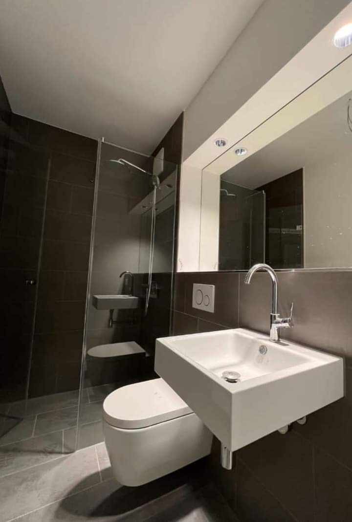 Nowoczesna łazienka z ciemnymi płytkami, wiszącą umywalką z chromowaną baterią, toaletą typu Rimless i kabiną prysznicową ze szklanymi drzwiami.