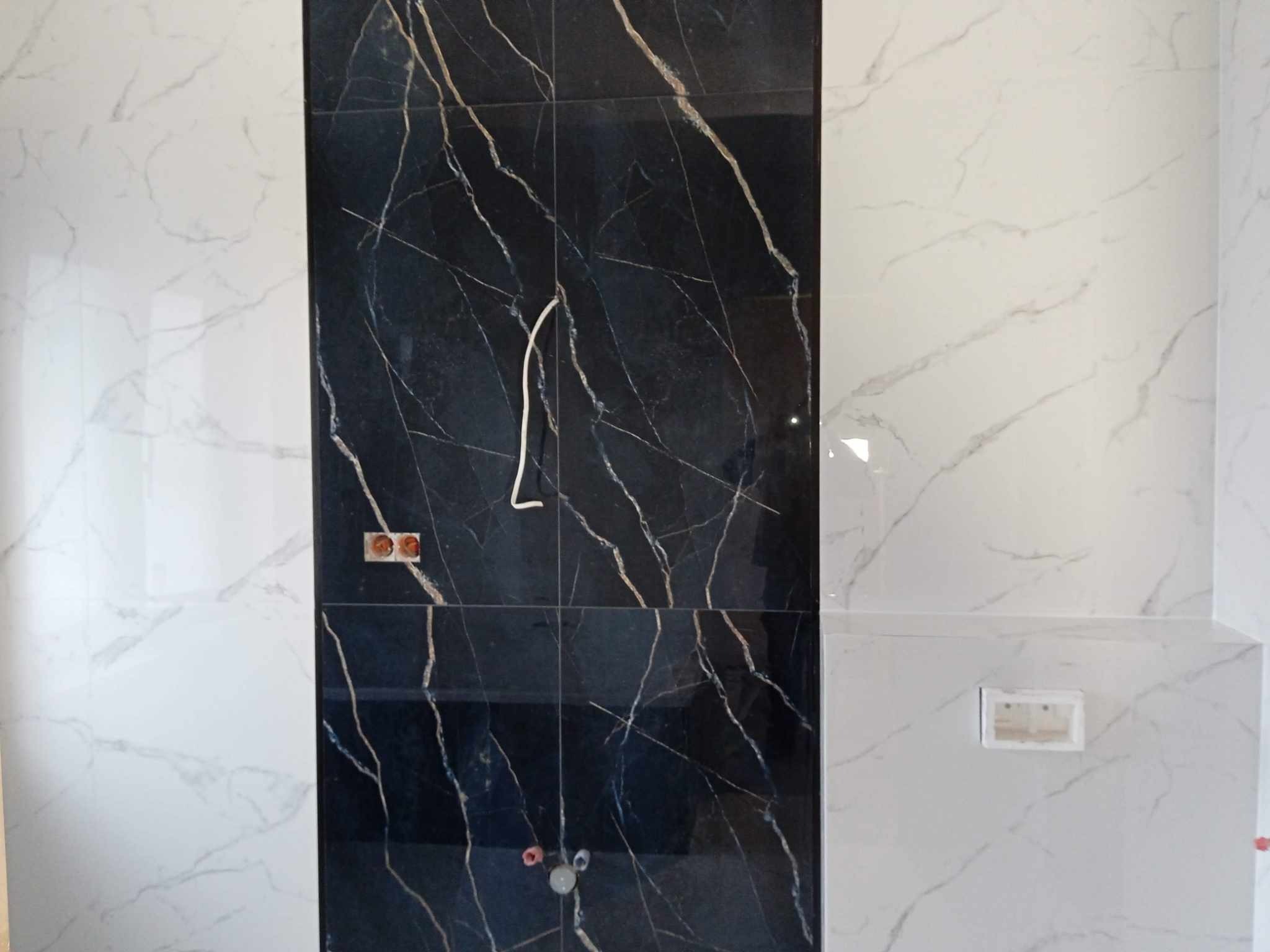 Ściana łazienki wyłożona płytkami w dwóch kolorach: czarnym z marmurowym wzorem i białym z szarymi żyłami, widoczne puszki elektryczne i wystające kable.