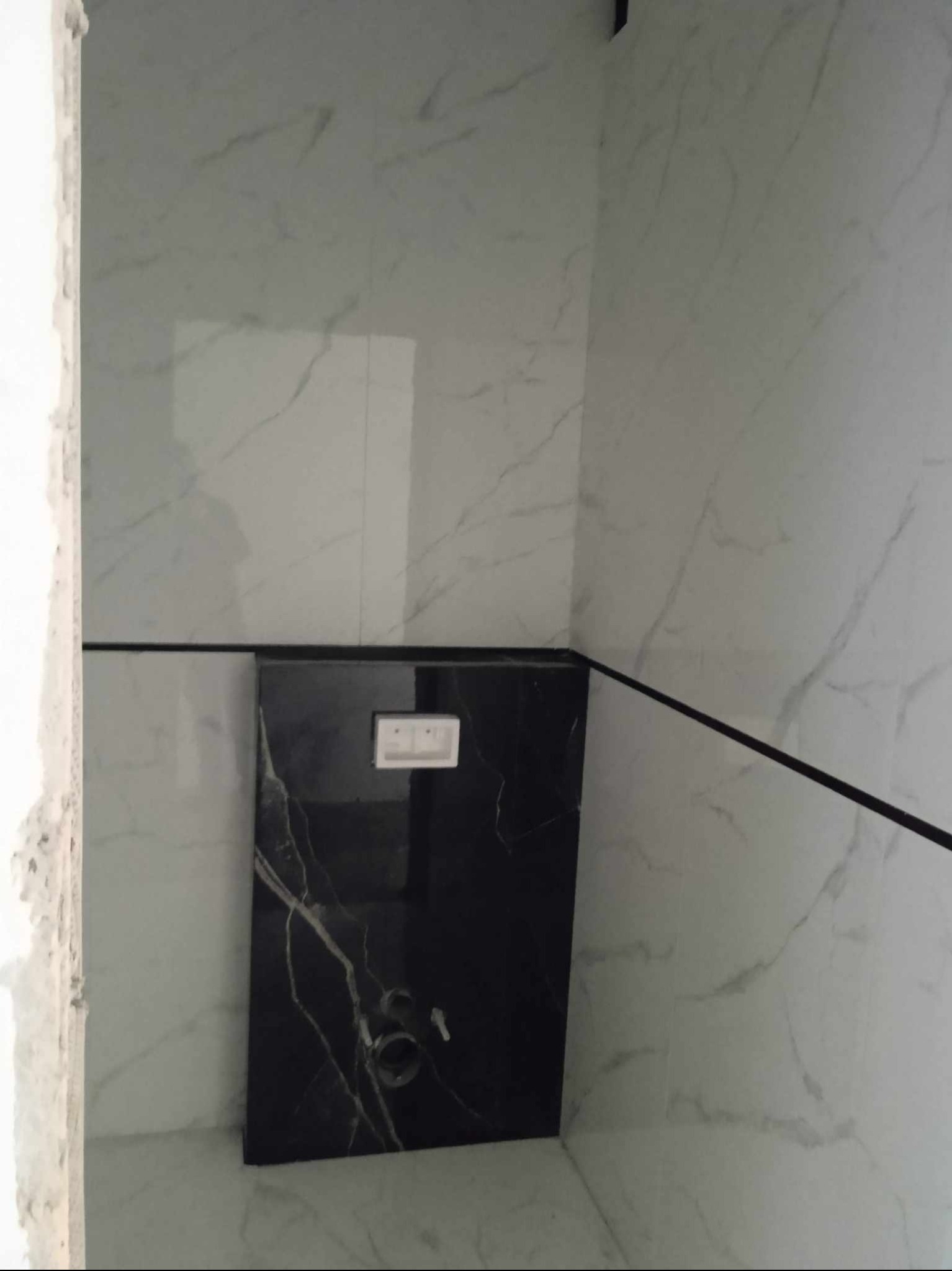 Fragment łazienki z płytkami imitującymi marmur, czarna zabudowa stelaża podtynkowego z białym przyciskiem spłukującym oraz czarny pasek dekoracyjny między płytkami.