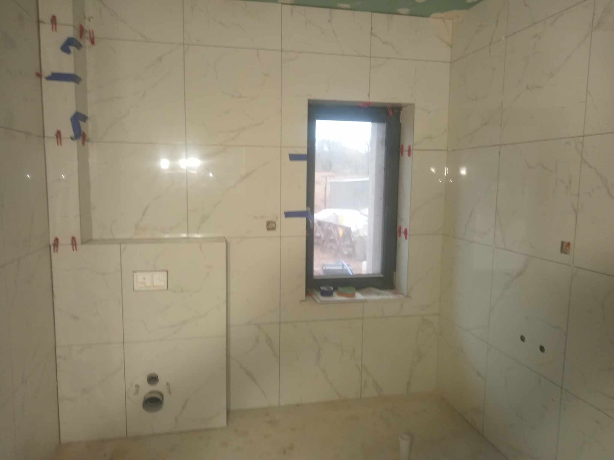 Ściany łazienki wyłożone białymi płytkami imitującymi marmur, z widocznymi krzyżykami dystansowymi i taśmą malarską, okno z ciemną ramą, podtynkowa spłuczka WC z przyciskami i otworem na muszlę.