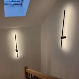 Dwa nowoczesne, czarne kinkiety LED na białej ścianie klatki schodowej, oświetlające przestrzeń w górę i w dół, z widocznym oknem dachowym w tle.