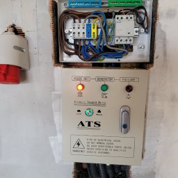 TK - INSTALATORSTWO ELEKTRYCZNE Tomasz Koprek - Skrzynka elektryczna ATS (Automatic Transfer Switch) z widocznymi przewodami i elementami sterującymi, zamontowana na surowej ścianie z widoczną siatką wzmacniającą.
