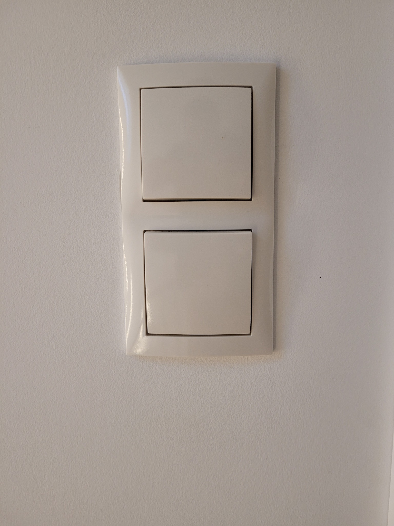 Biały, podwójny włącznik światła zamontowany na gładkiej, białej ścianie. Widoczny delikatny połysk plastiku.