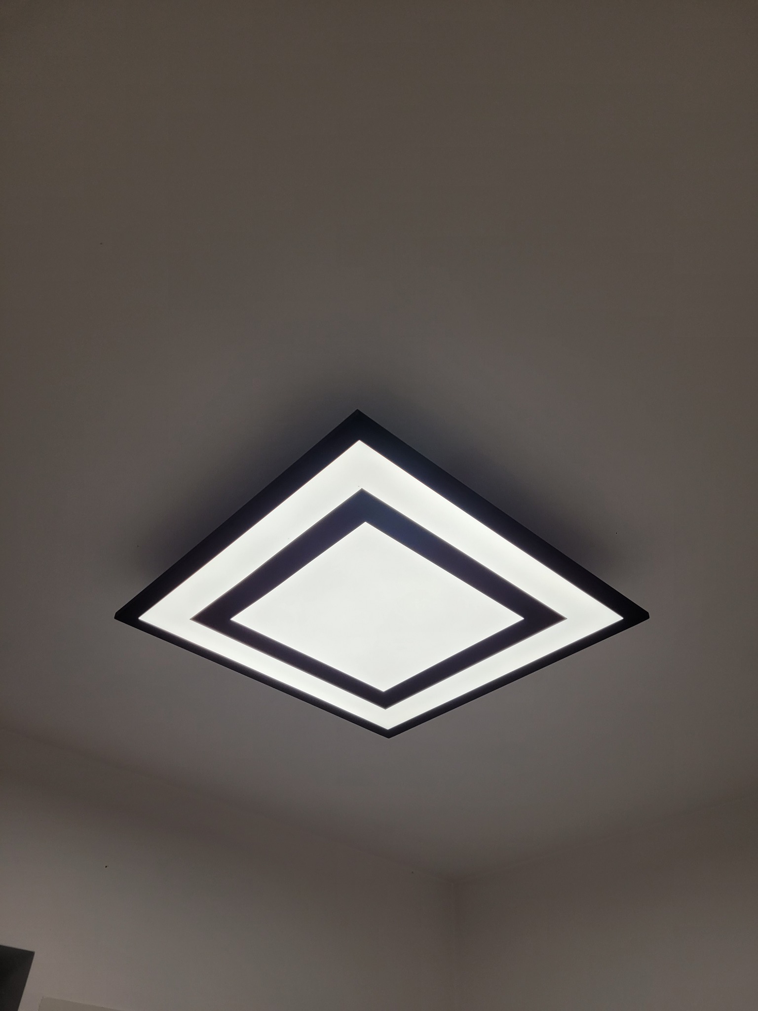 Nowoczesna, kwadratowa lampa sufitowa LED z czarną ramą, emitująca jasne, równomierne światło, zamontowana na białym suficie pod kątem od dołu.