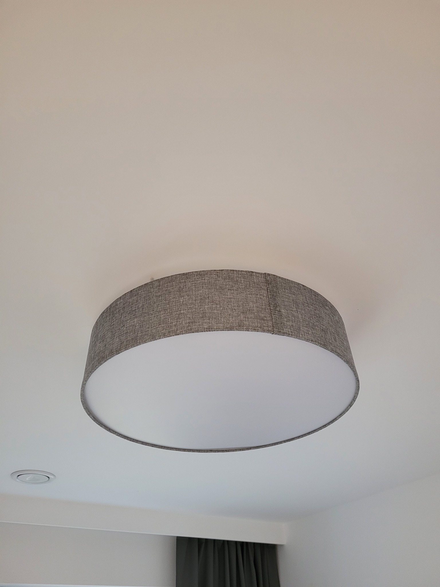 Szare, okrągłe oświetlenie sufitowe z tekstylnym abażurem, widziane pod kątem, z widocznym fragmentem sufitu i drugiego, mniejszego oświetlenia w tle.