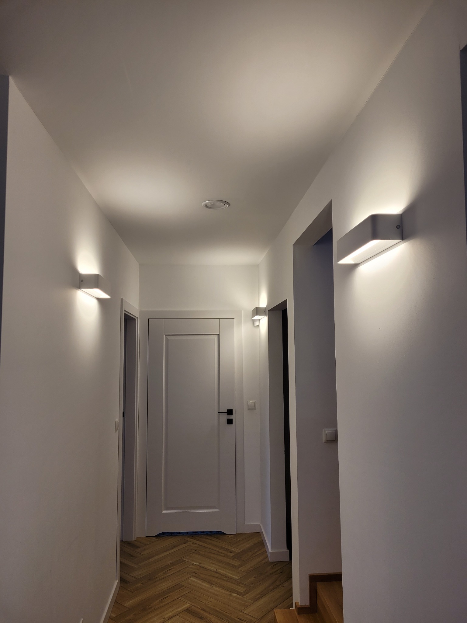 Korytarz z białymi ścianami i parkietem w jodełkę, oświetlony nowoczesnymi, prostokątnymi lampami ściennymi z ciepłym światłem.