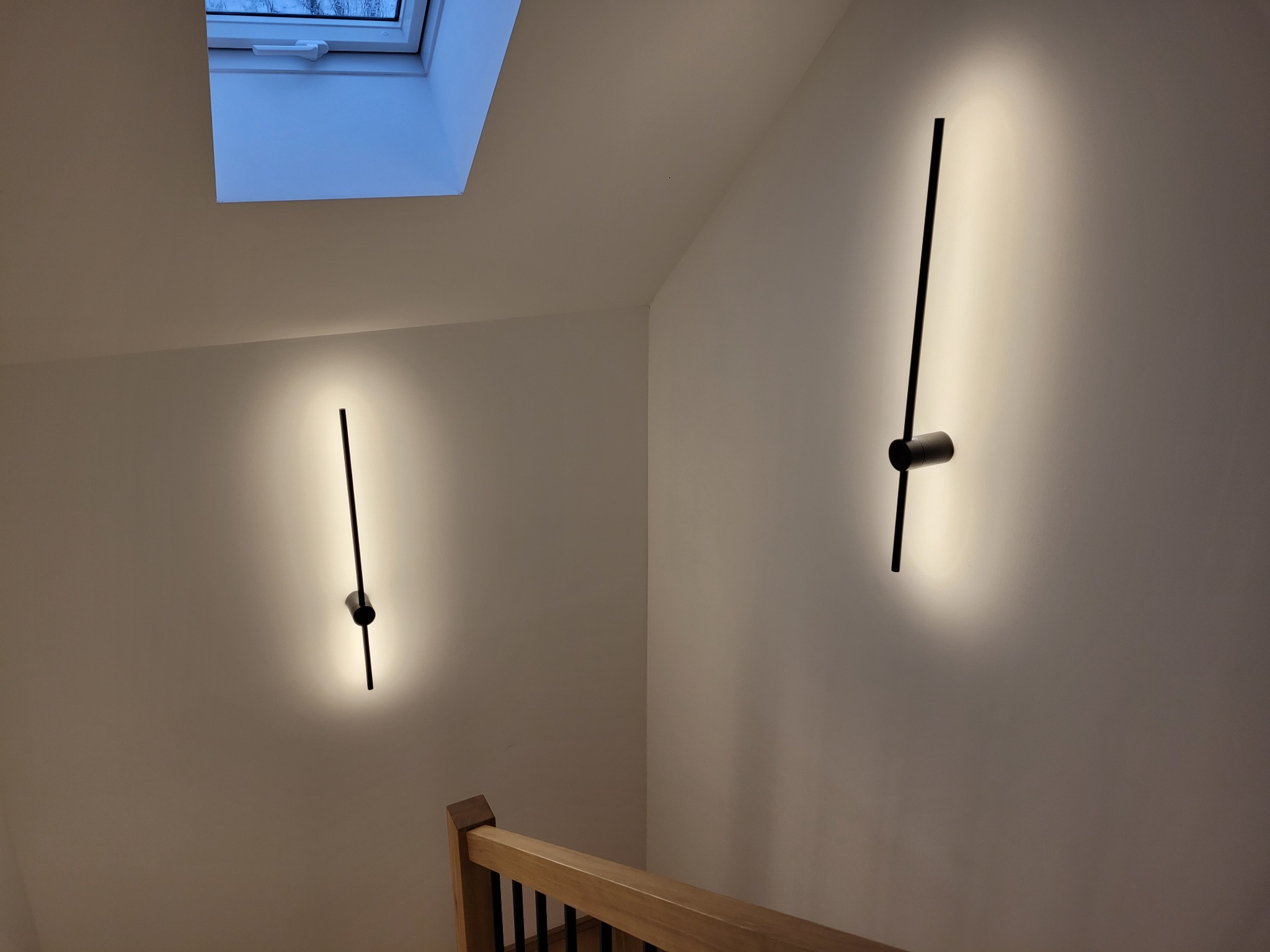 Dwa nowoczesne, czarne kinkiety LED na białej ścianie klatki schodowej, oświetlające przestrzeń w górę i w dół, z widocznym oknem dachowym w tle.