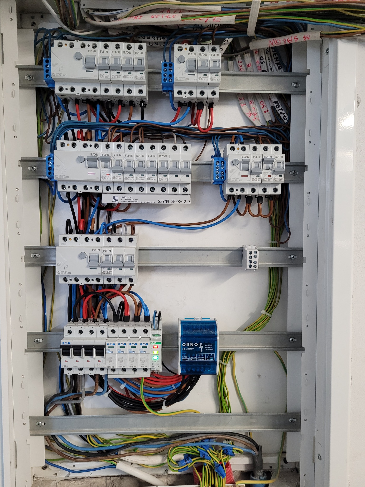 Rozdzielnia elektryczna z bezpiecznikami Eaton i ORNO, z widocznymi przewodami w różnych kolorach. Instalacja w białej obudowie z szynami montażowymi.