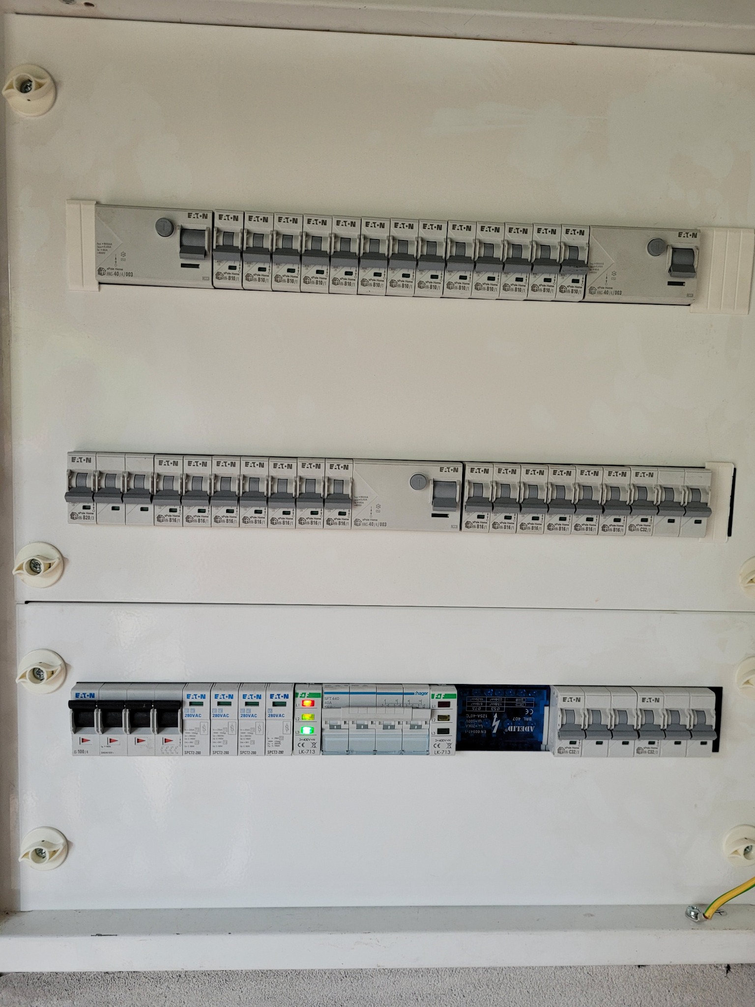 Biała skrzynka rozdzielcza z trzema rzędami bezpieczników marki Eaton i Hager, widoczne szczegóły instalacji elektrycznej wewnątrz budynku.