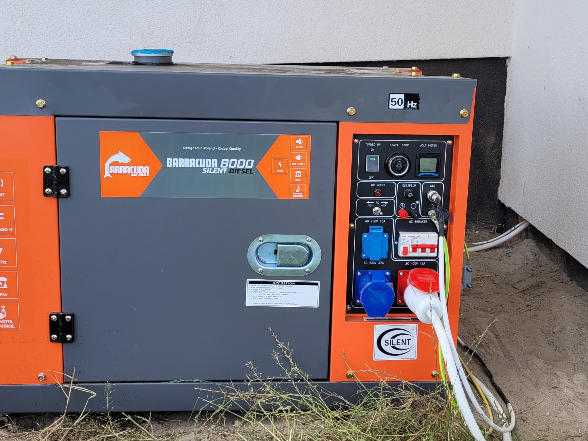 Agregat prądotwórczy Barracuda 8000 Silent Diesel, z widocznym panelem sterowania i podłączonymi kablami, stoi przy ścianie budynku.