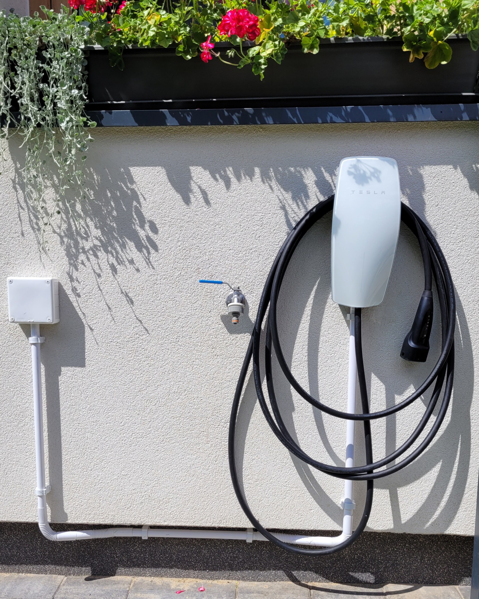 Biała stacja ładowania Tesla Wall Connector Gen 3 zamontowana na elewacji budynku, z kablem i puszką elektryczną. Nad stacją widoczne kwiaty w donicy.