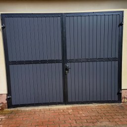 RAJA-TECHNIK - Zamknięta, dwuskrzydłowa brama garażowa z szarymi panelami pionowymi i czarną ramą, zabezpieczona zamkiem na tle otynkowanej ściany i czerwonej kostki brukowej.