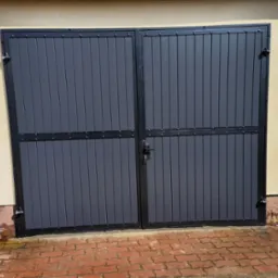 Zamknięta, dwuskrzydłowa brama garażowa z szarymi panelami pionowymi i czarną ramą, zabezpieczona zamkiem na tle otynkowanej ściany i czerwonej kostki brukowej.