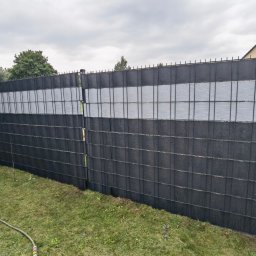 RAJA-TECHNIK - Nowoczesne ogrodzenie panelowe z poziomymi pasami w kolorze czarnym i szarym, widok z zewnątrz na tle zielonego trawnika i pochmurnego nieba, z taczką ogrodową w lewym dolnym rogu.