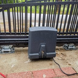 RAJA-TECHNIK - Czarny automat do bramy przesuwnej, widoczne krople deszczu, klucz w zamku, szyna bramy i kostka brukowa.