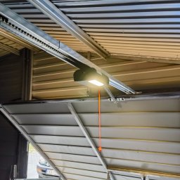 RAJA-TECHNIK - Wnętrze garażu z widocznym mechanizmem napędu bramy garażowej, łańcuch, silnik z oświetleniem i częściowo otwarta brama segmentowa.