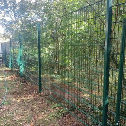 RAJA-TECHNIK - Zielone ogrodzenie panelowe z widocznym słupkiem i siatką na tle drzew i ziemi, z pozostawionym wężem ogrodowym.