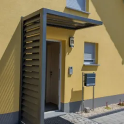 Nowoczesne zadaszenie nad wejściem do domu z lamelami, skrzynka na listy i elewacja w kolorze żółtym, widok na podjazd i auto.