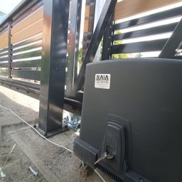 RAJA-TECHNIK - Zasuwna brama z drewnianymi elementami i czarną metalową ramą, widoczny automat z kluczykiem w zamku, kabel zasilający i podłoże z kostki brukowej.