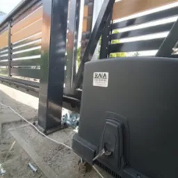 Zasuwna brama z drewnianymi elementami i czarną metalową ramą, widoczny automat z kluczykiem w zamku, kabel zasilający i podłoże z kostki brukowej.