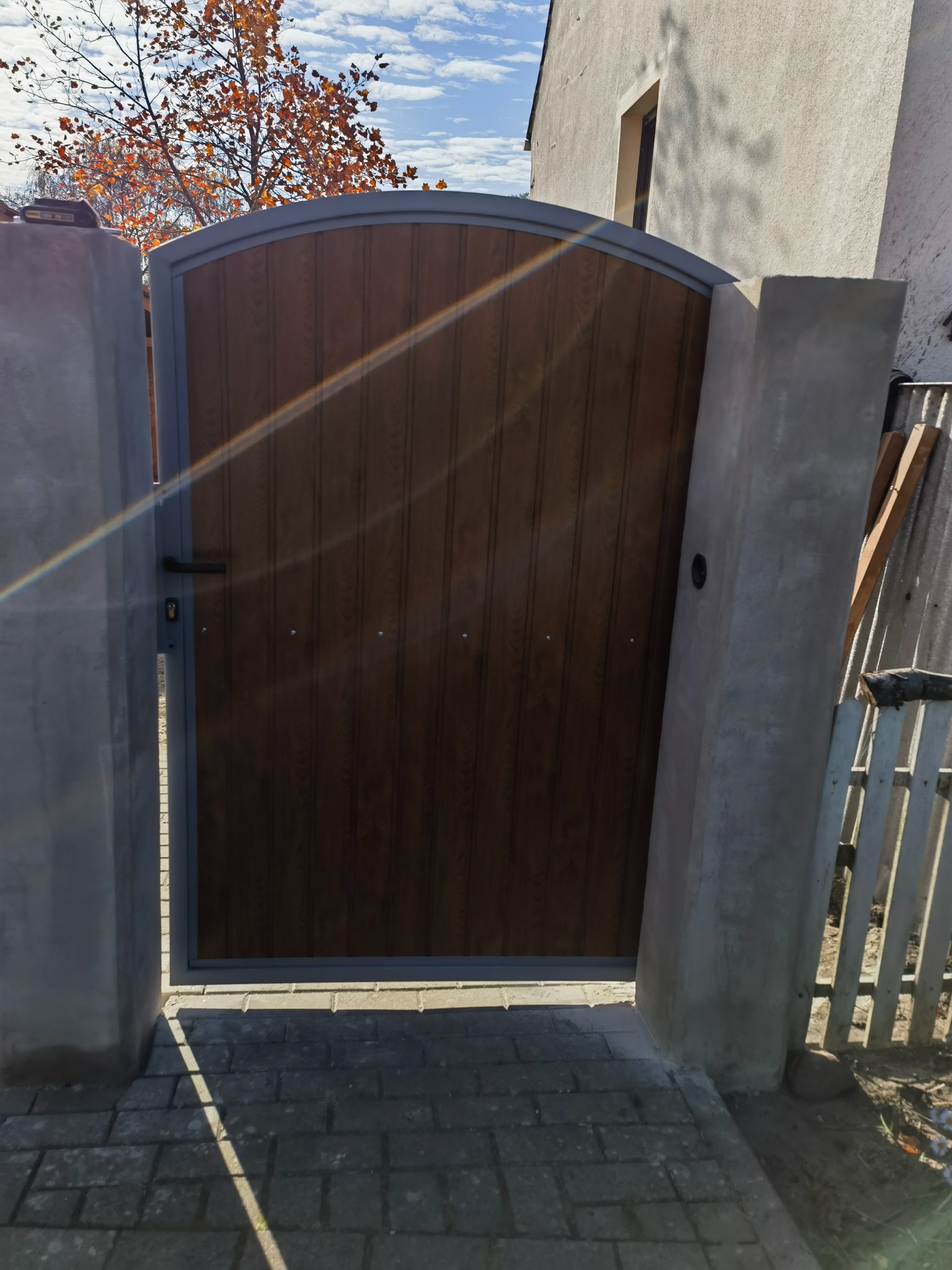 Zamknięta brama wejściowa z drewnopodobnym wypełnieniem i szarą ramą, osadzona między betonowymi słupkami, na tle domu i drzewa z pomarańczowymi liśćmi.