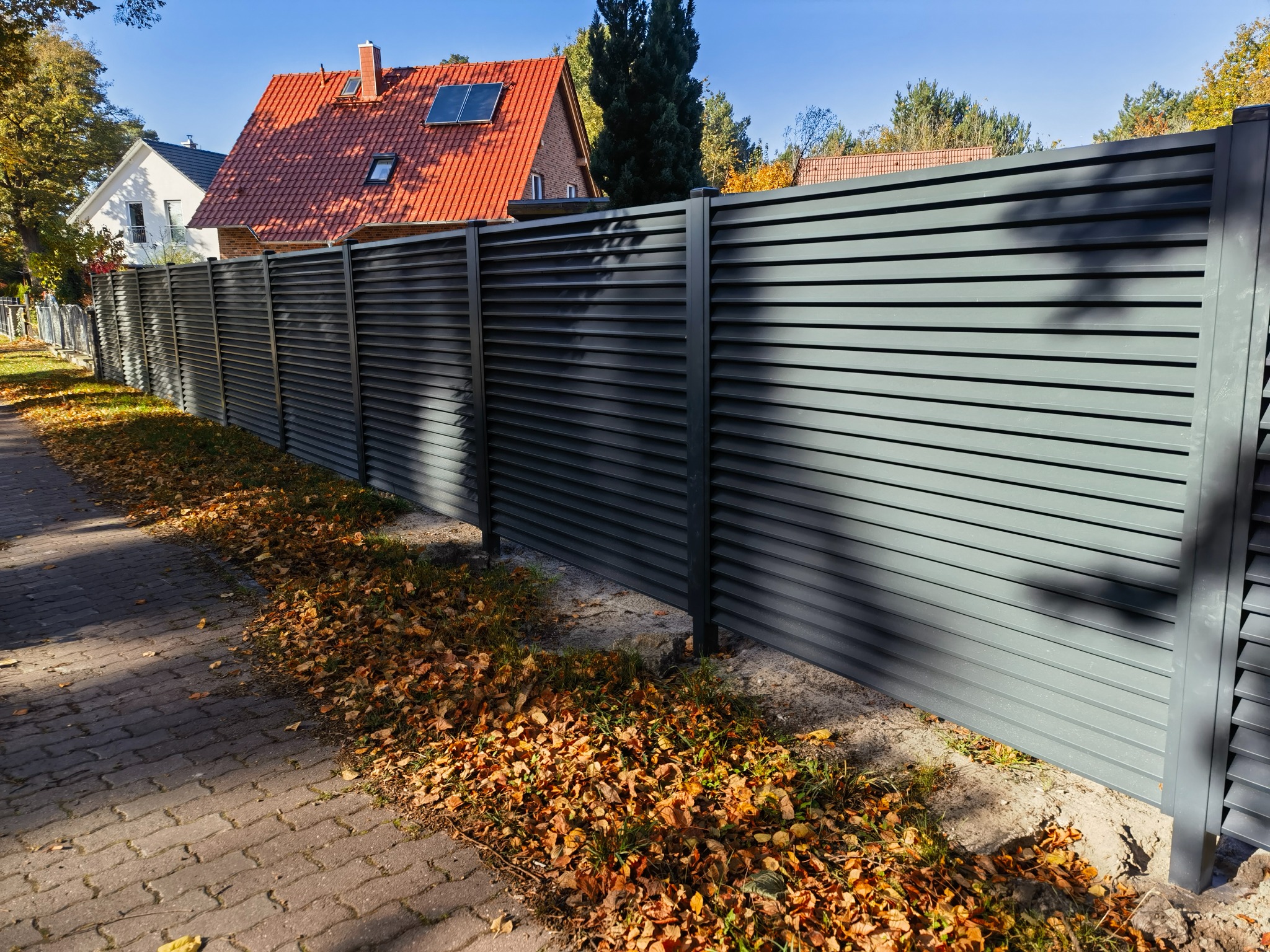 Nowoczesne ogrodzenie metalowe panelowe w kolorze grafitowym, z poziomymi lamelami, oddzielające posesję od chodnika z kostki, jesienne liście na trawie u podnóża ogrodzenia, dom z czerwoną...
