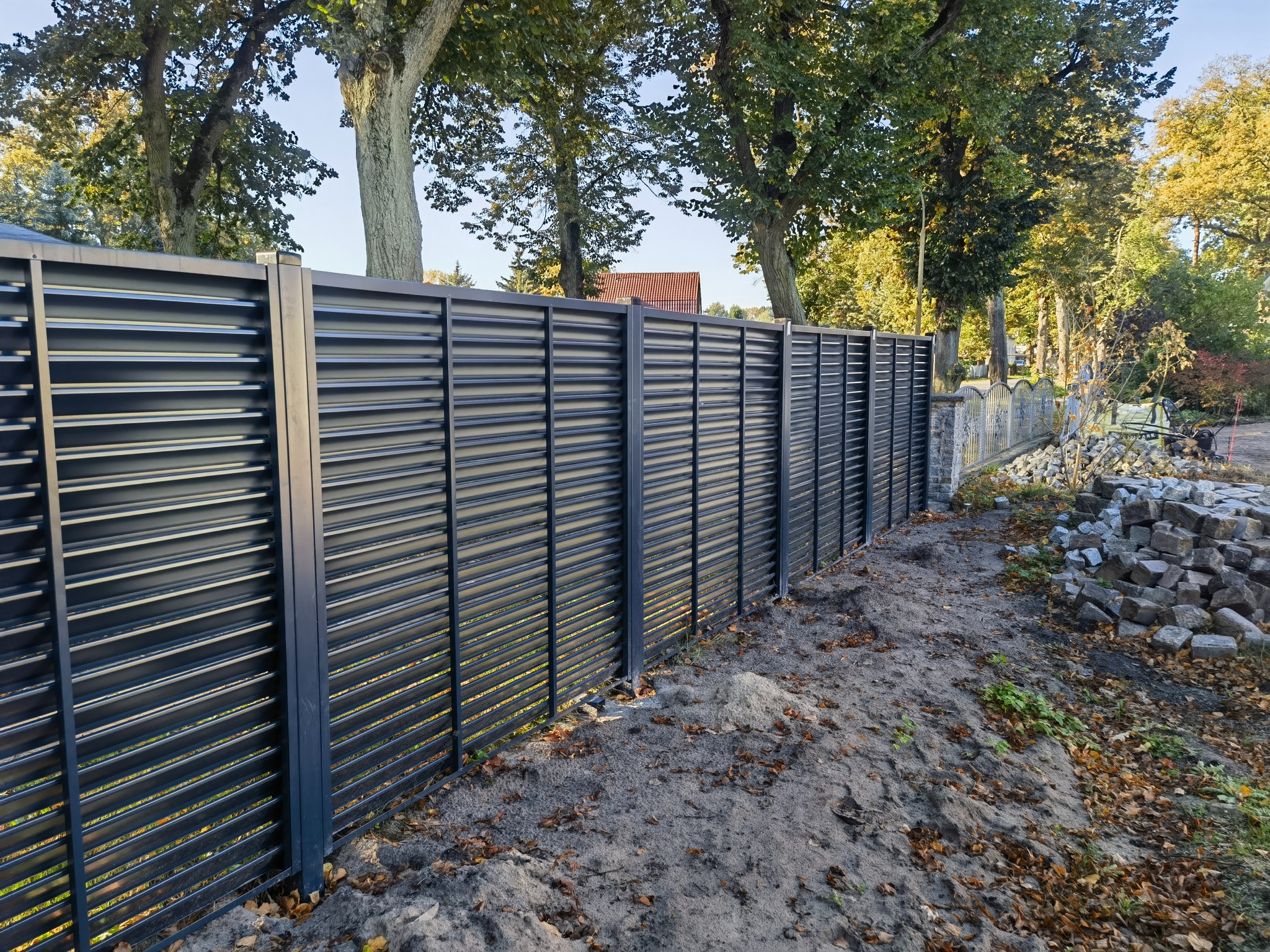 Długi, nowoczesny, poziomy, szary płot lamelowy z metalu, widok z boku, z ziemią i liśćmi u podstawy, drzewa w tle, część starego kamiennego muru po prawej stronie, stos kamieni obok.