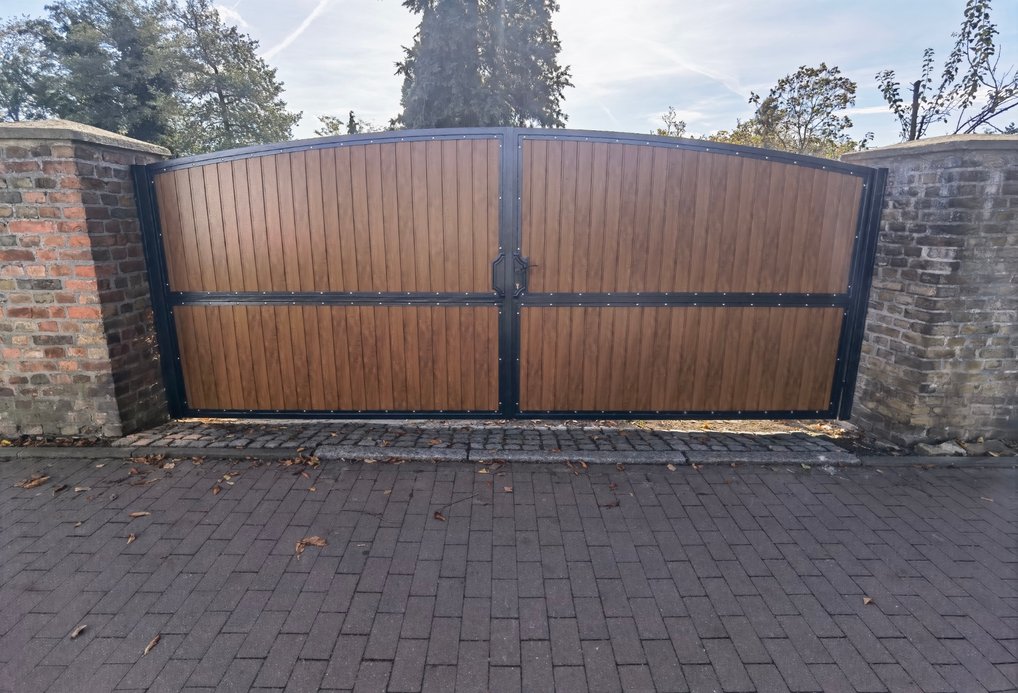 Dwuskrzydłowa brama wjazdowa z drewnopodobnym wypełnieniem, osadzona między murowanymi filarami z cegły, widoczna z poziomu brukowanej drogi.