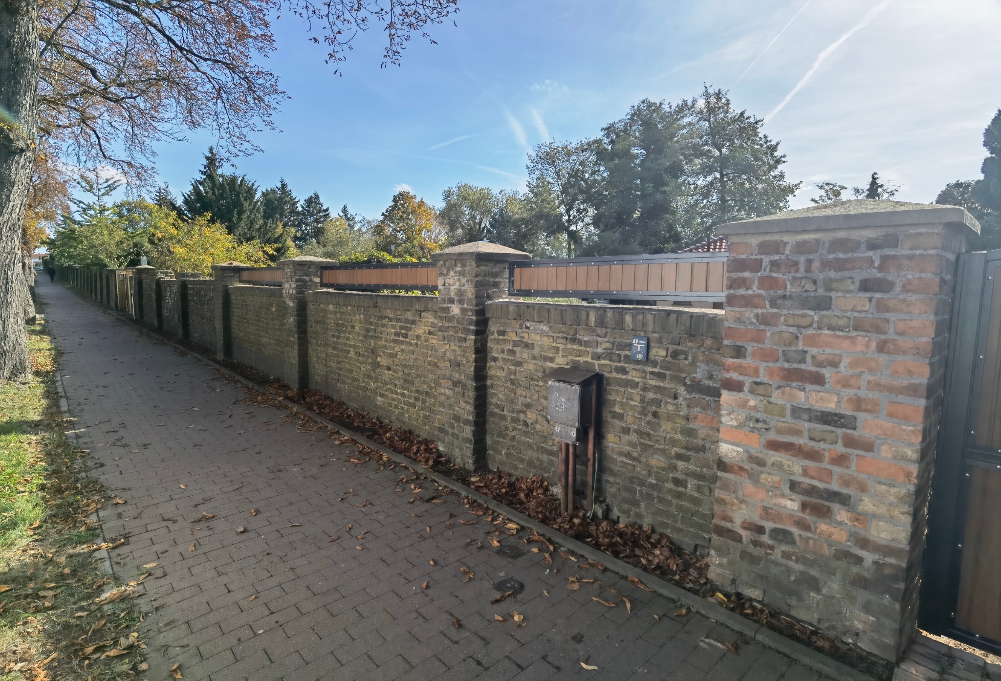 Długi, murowany płot z cegły z drewnianymi przęsłami i metalową bramą, widziany z perspektywy chodnika w słoneczny dzień.