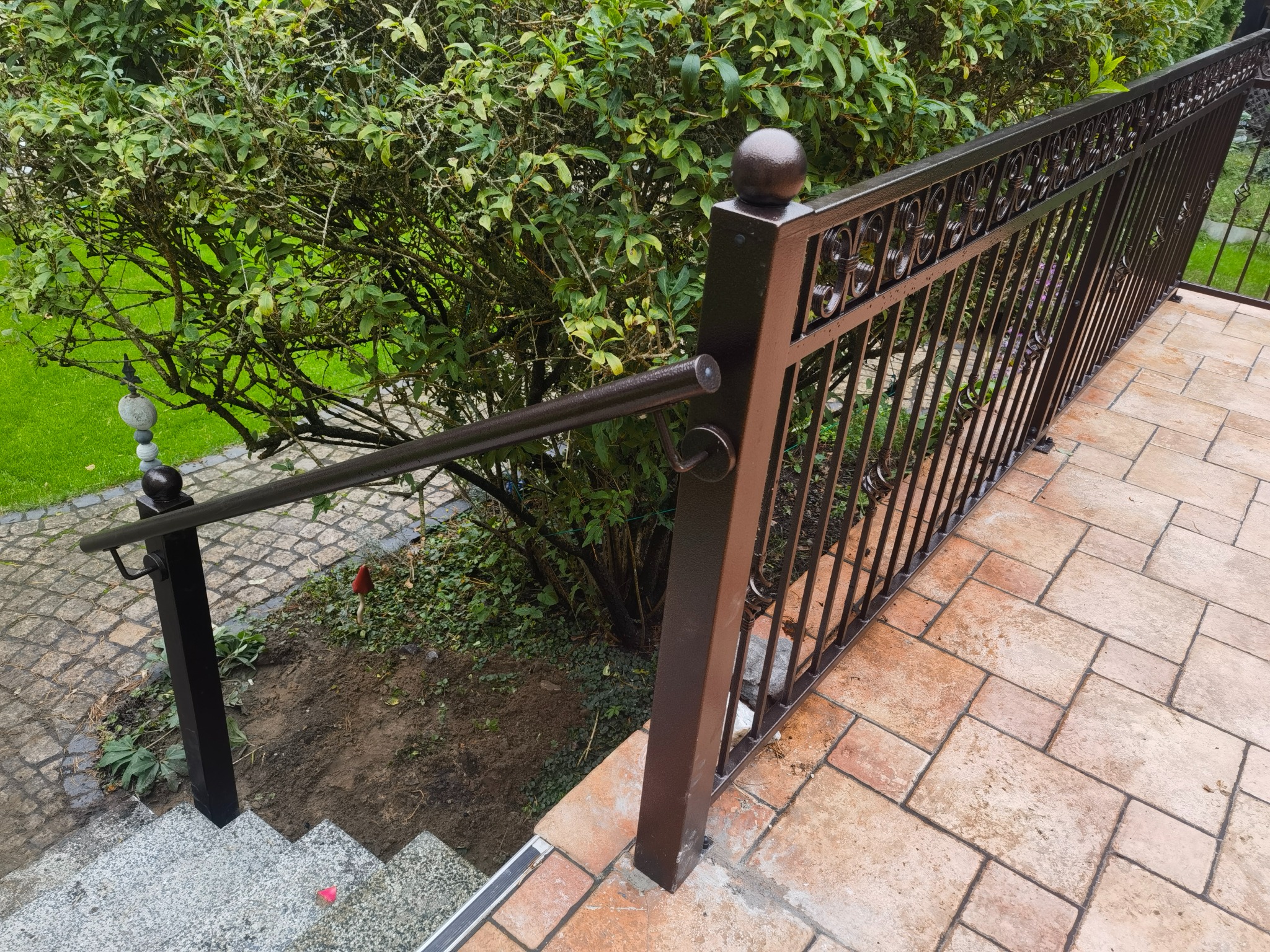 Metalowa balustrada schodowa w kolorze ciemnego brązu z ozdobnymi elementami, prowadząca w dół po schodach wyłożonych szarym kamieniem, otoczona zielenią.