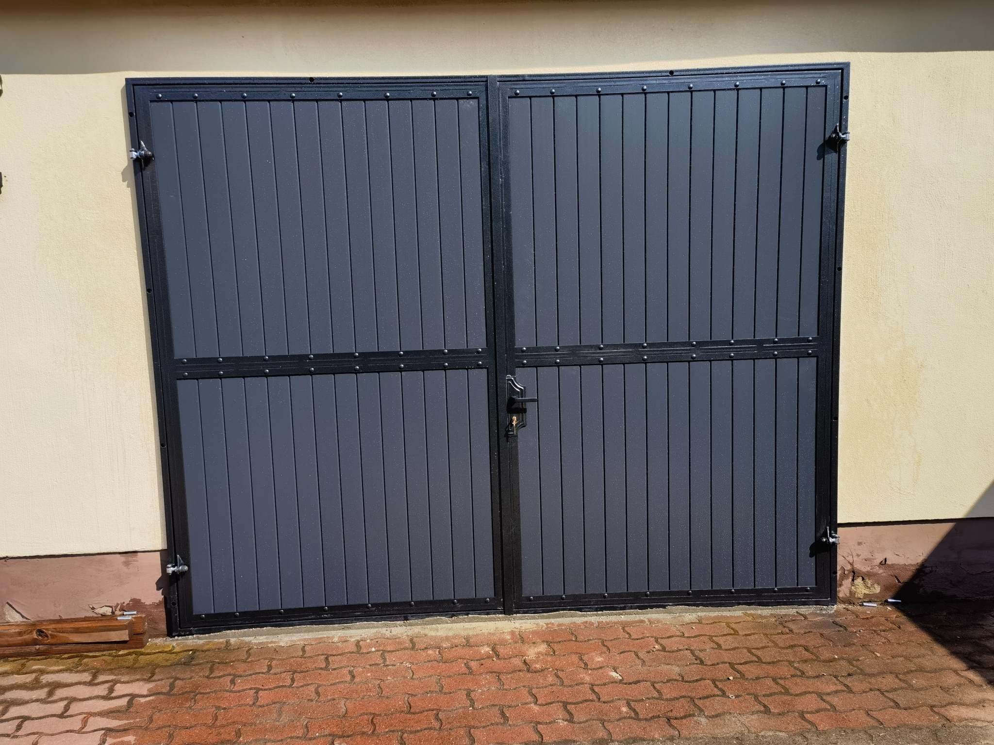 Zamknięta, dwuskrzydłowa brama garażowa z szarymi panelami pionowymi i czarną ramą, zabezpieczona zamkiem na tle otynkowanej ściany i czerwonej kostki brukowej.