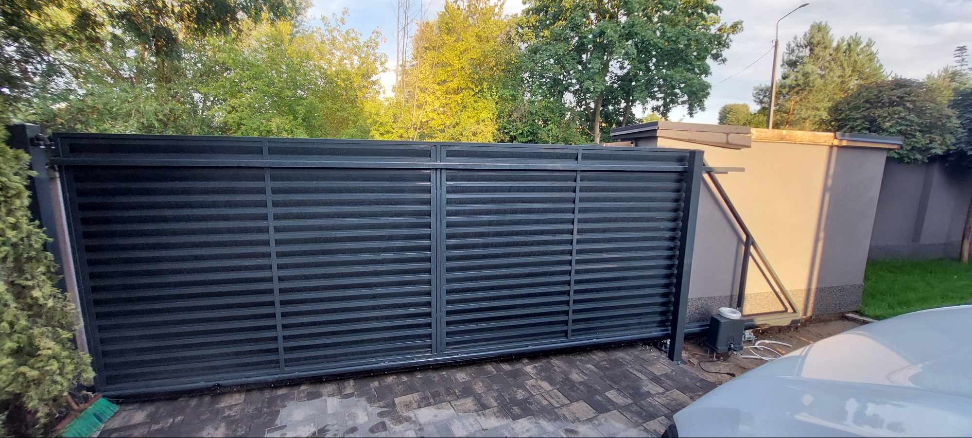 Nowoczesna, przesuwna brama w kolorze grafitowym z poziomymi panelami, zamontowana na posesji z kostką brukową i zielenią w tle, widoczny napęd elektryczny.